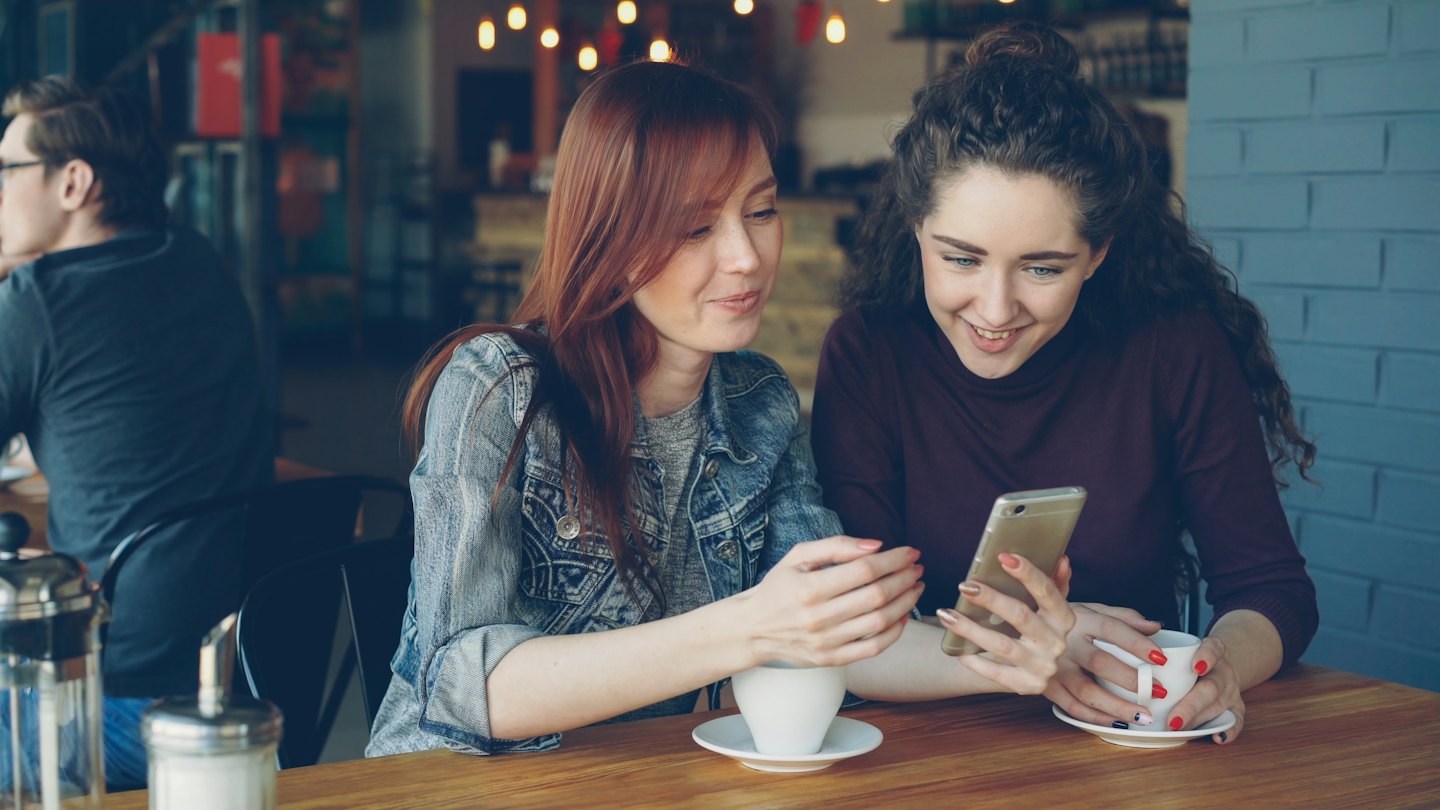 Two women looking at a smartphone at a cafe table.